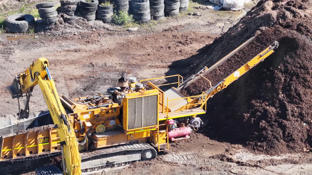 An excavator transfers mulch into a processing machine in a dirt-filled industrial site under bright daylight