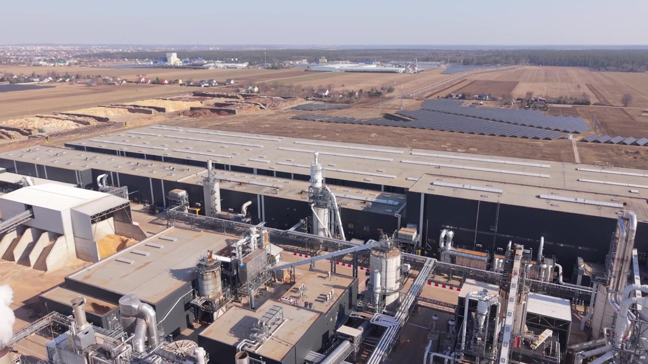 Campus industriale aereo con pannelli solari sul tetto e lungo magazzino, camion parcheggiati presso le aree di carico, campi visibili sullo sfondo, ampia prospettiva paesaggistica per energia e logistica