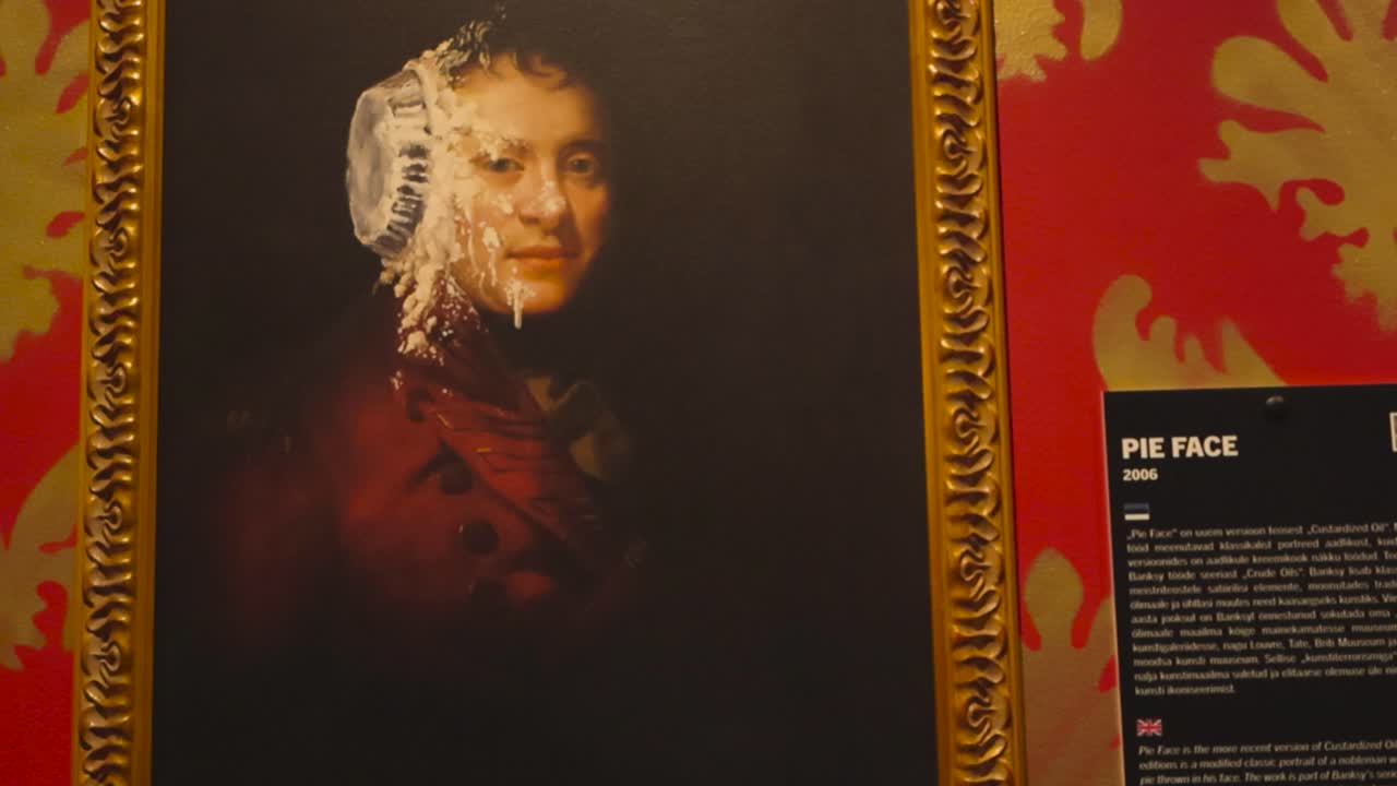 Painting of musician Bach being hit in the face with a pie called Pie Face done by Banksy on display at his exhibition at Telliskivi. Funny painting is on a red and golden wall framed beautifully.