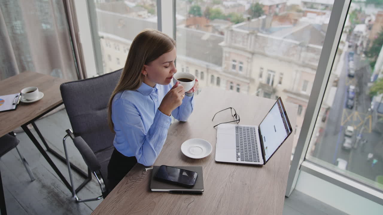 Blonde female sitting at desk in office holding a cup of coffee. Calm relaxed lady enjoying lunch time at work place. High angle view.