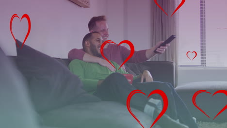 animation of heart icons over diverse gay couple smiling and watching tv