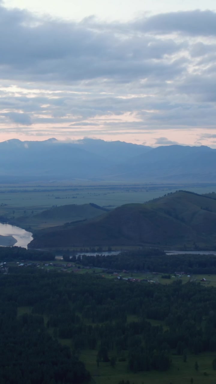 Weitwinkel-Landschaftsaufnahme von Zamul'ta, Altai-Republik, Russland, mit bewaldeten Hügeln, Feldern wie ein Flickenteppich und fernen Bergen unter dramatisch bewölktem Himmel.