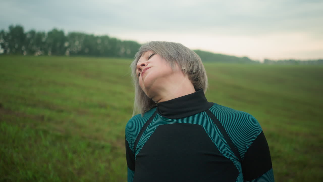 검은색과 초록색 의상을 입은 여성, 눈을 감고, 조용한 야외 환경에서 머리를 오른쪽에서 왼쪽으로 부드럽게 기울이고, 긴장을 풀고 신체 의식을 촉진하기 위해 주의 깊은 목 스트레칭을 연습합니다.