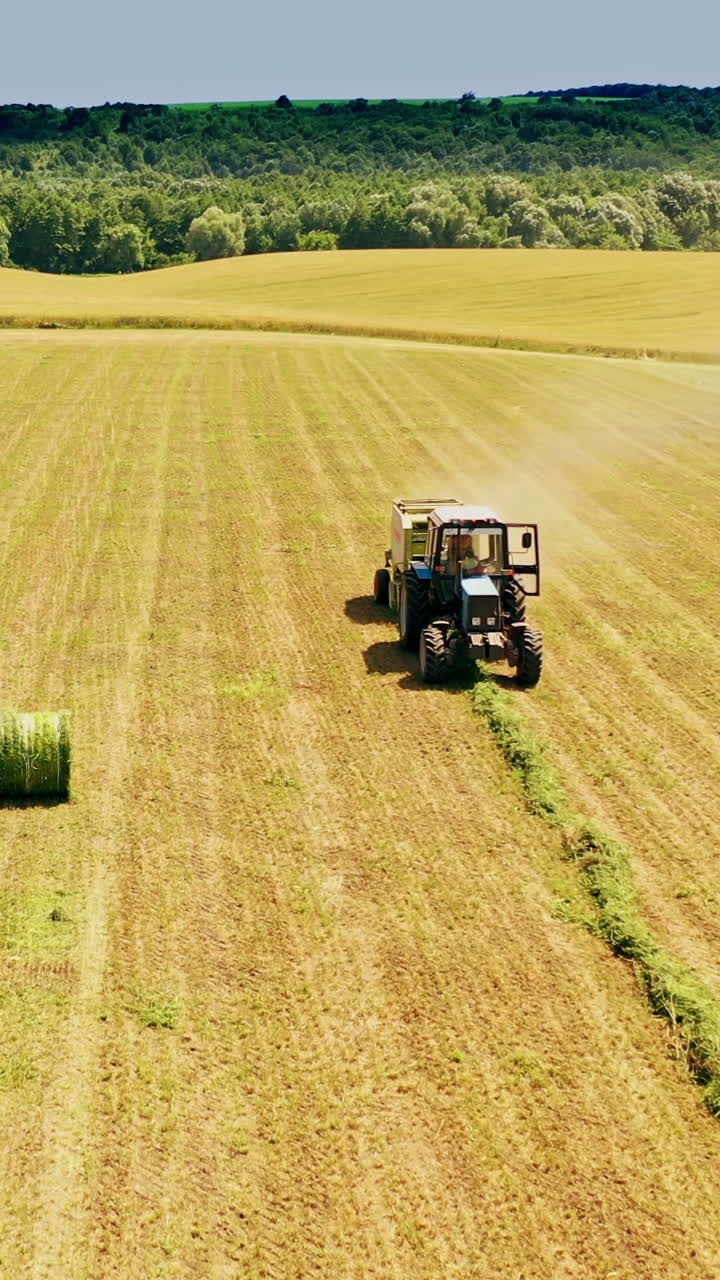 Tractor at work and some bales on the field on natural background. Agricultural tractor in field pressing grass into round bundles at sunny summer day. Aerial view. Vertical video