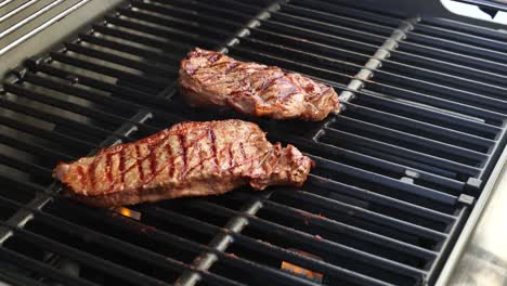 two beef steaks being grilled on a barbecue, with tongs used to flip them. bright outdoor lighting enhances the scene