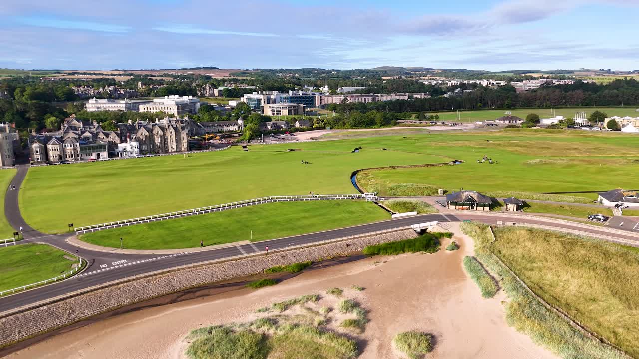 Drone footage glides above green fairways, sandy dunes, and Victorian brick buildings at a famous golf course in St Andrews, Scotland, under bright daylight