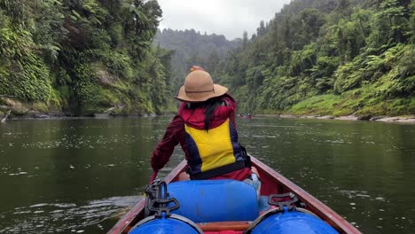 A-woman-with-black-hair-wearing-a-sun-hat,-red-jacket,-and-yellow-life-vest-paddles-down-the-Whanganui-River-in-a-canoe-in-a-lush-forested-river-valley