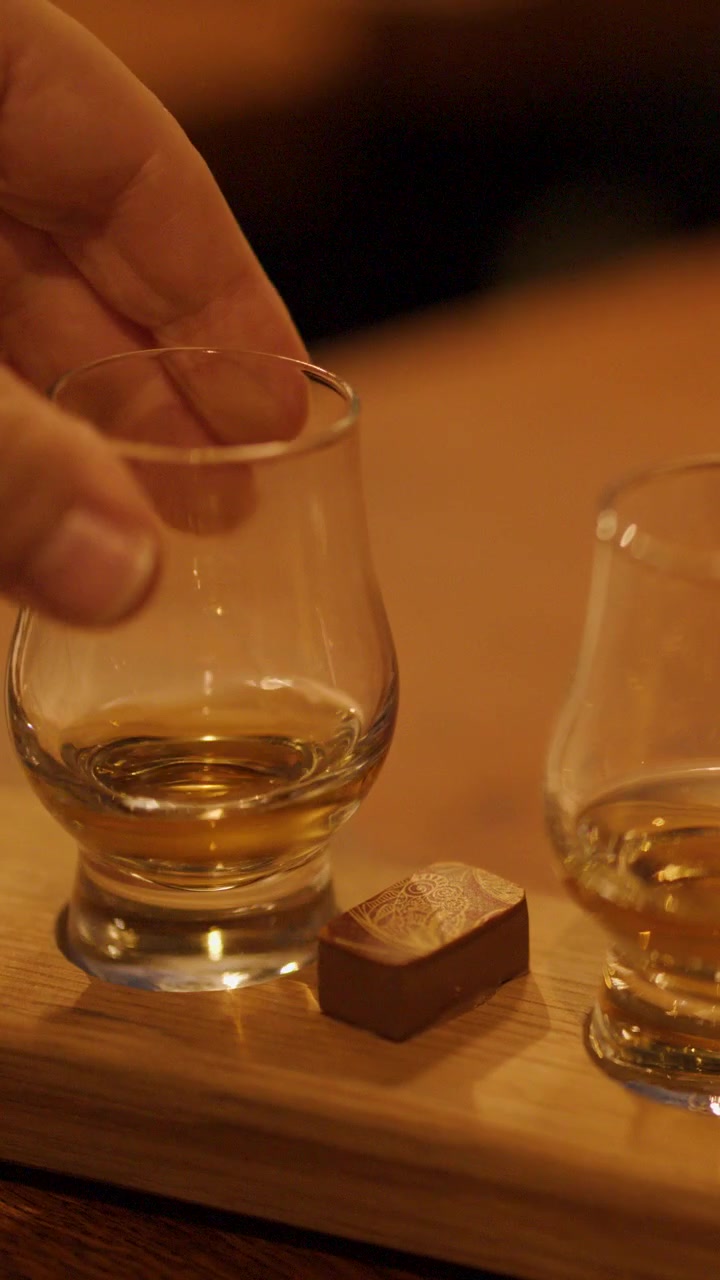 Una mano solleva un bicchiere di whisky da un vassoio di legno con cioccolato, illuminazione calda, atmosfera interna accogliente