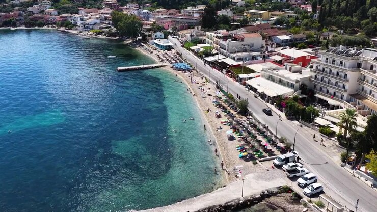 Vista aérea da praia de Benitses em Corfu, Grécia. Mar Jônico turquesa, espreguiçadeiras, estrada costeira com carros e resorts de verão ao longo da pitoresca costa mediterrânea