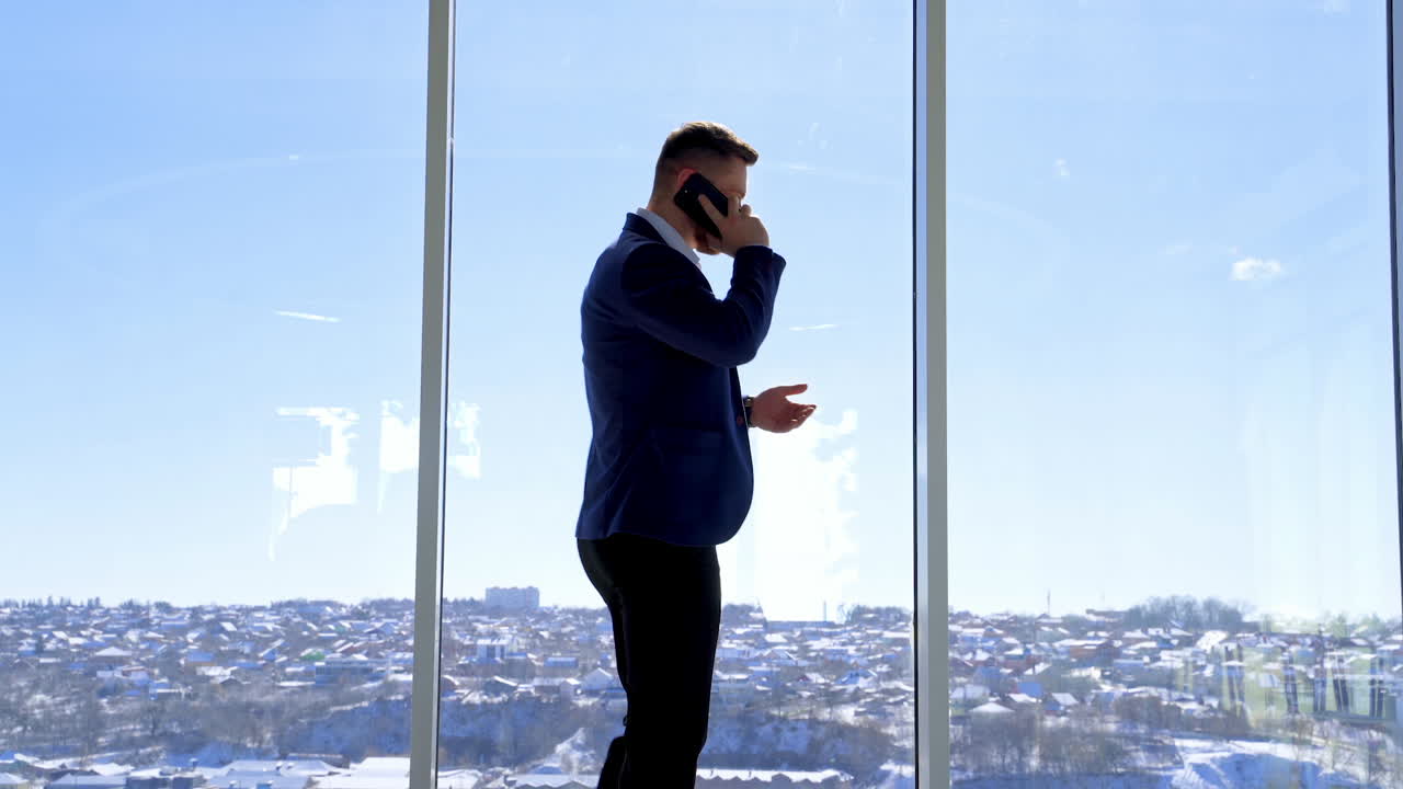 Businessman having a conversation through the phone. Young man in elegant suit standing near the large office window and talking the phone. Rear view.
