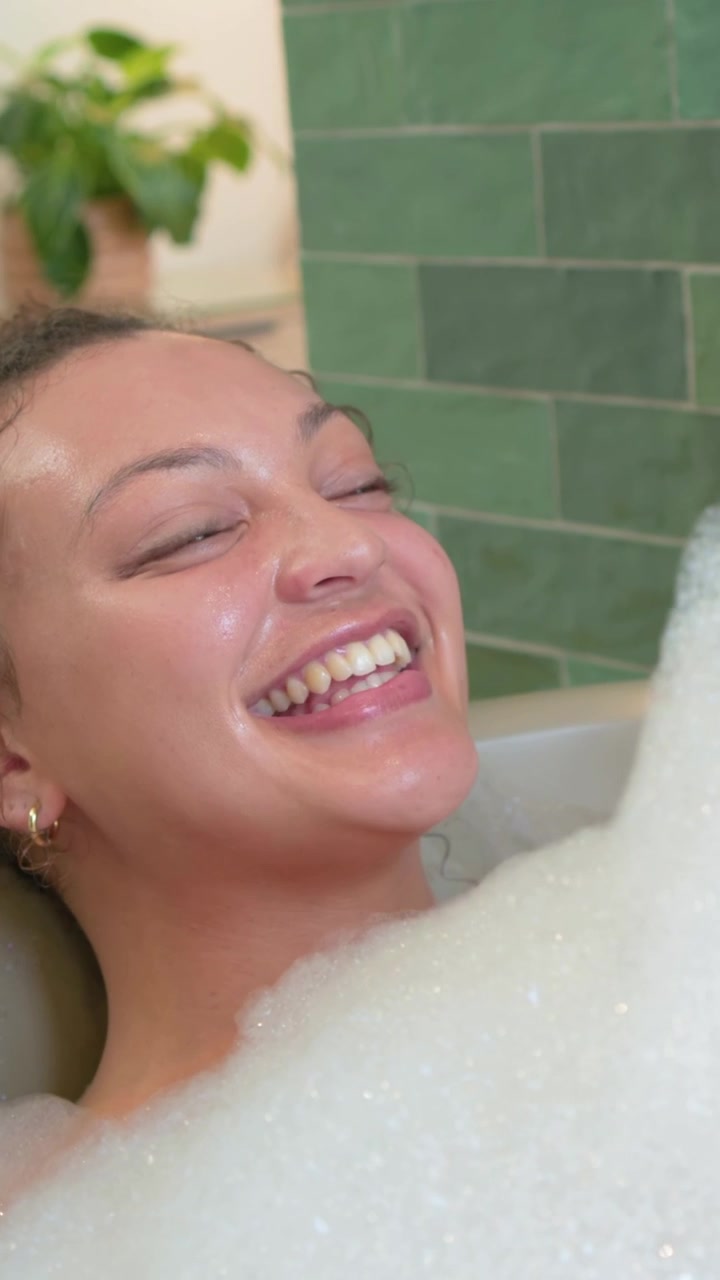 Vertical video: Blowing foam after faucet releasing foam, woman in bathtub scooping and relaxing