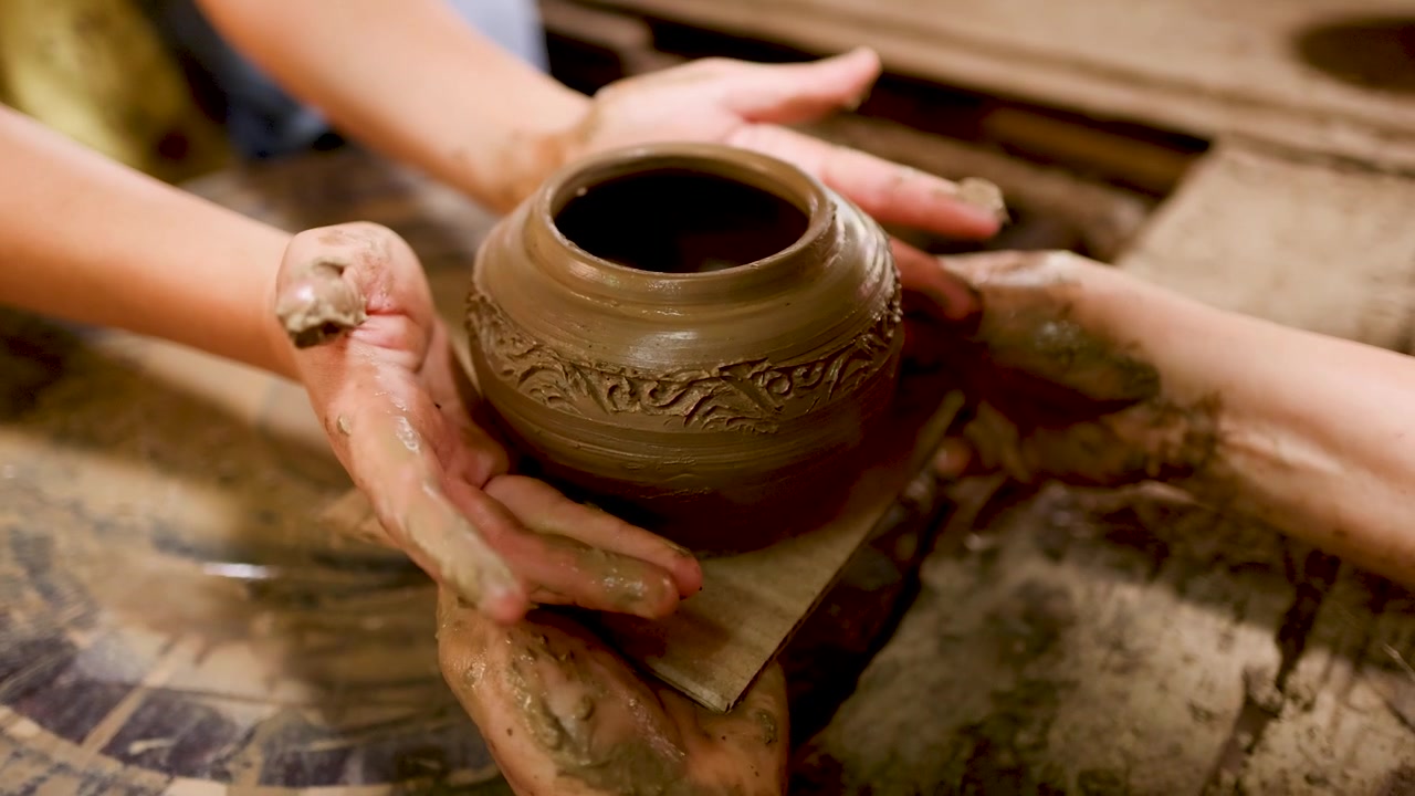 Primo piano delle mani di un artigiano che sollevano un vaso di ceramica dettagliato da un tornio da vasaio che gira.