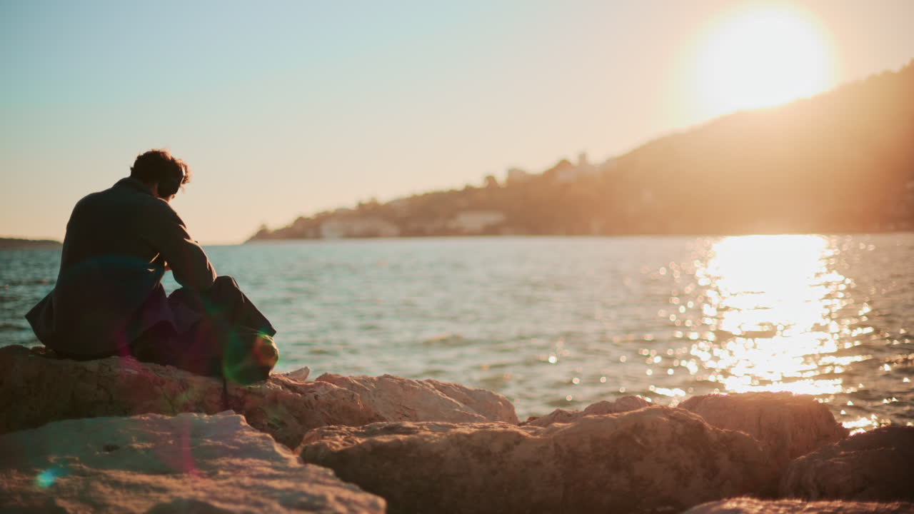 A man sitting alone on coastal rocks, looking down as the sun sets over the sea and hills, creating a warm contemplative mood