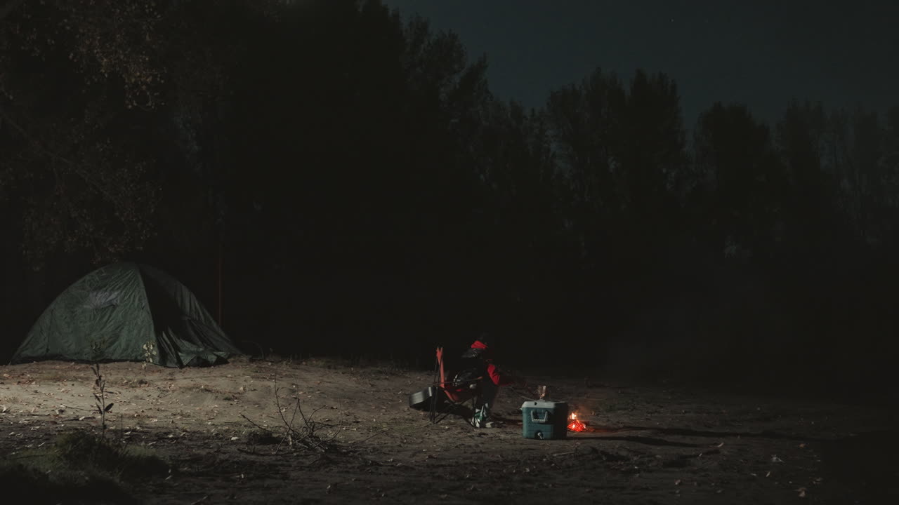 Stille lejrplads under stjernehimmel. Fredfyldt lejrplads med glødende bål og to hvilende figurer. Fredelig udendørs campingplads under nattehimlen med et svagt glødende bål og træer i silhuet