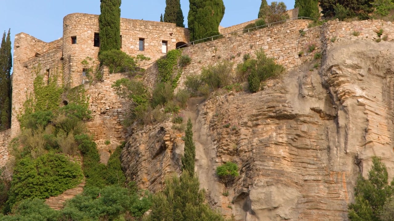 Panning shot of ancient fortress walls and cypress trees atop a rugged limestone cliffside.