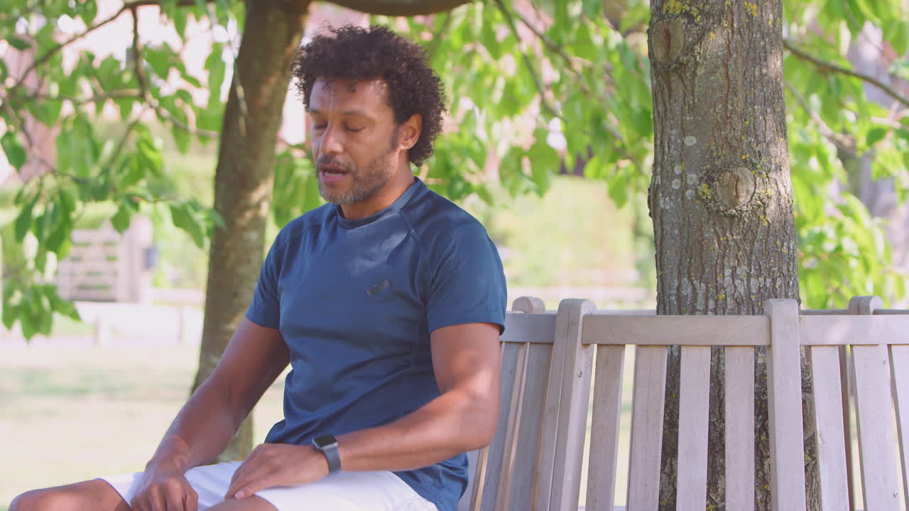 hombre maduro vestido con ropa de fitness se sienta en el asiento bajo el árbol monitoreando la actividad en el reloj inteligente