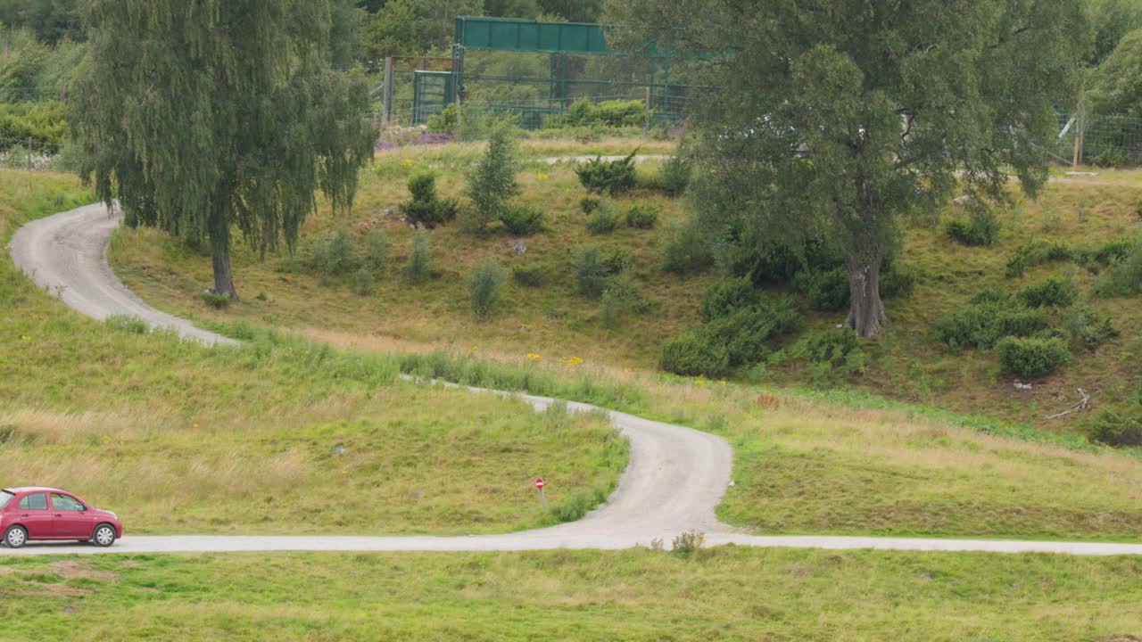 Red compact car travels on curving rural road through grassy Highlands landscape, daylight, static shot