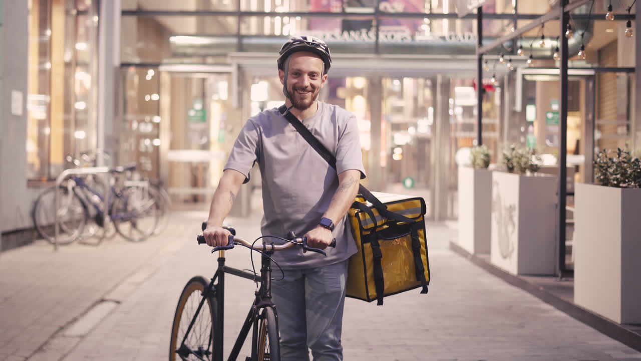 Happy Bearded Food Delivery Man Wearing A Thermal Backpack And Helmet Looks Directly Into The Camera