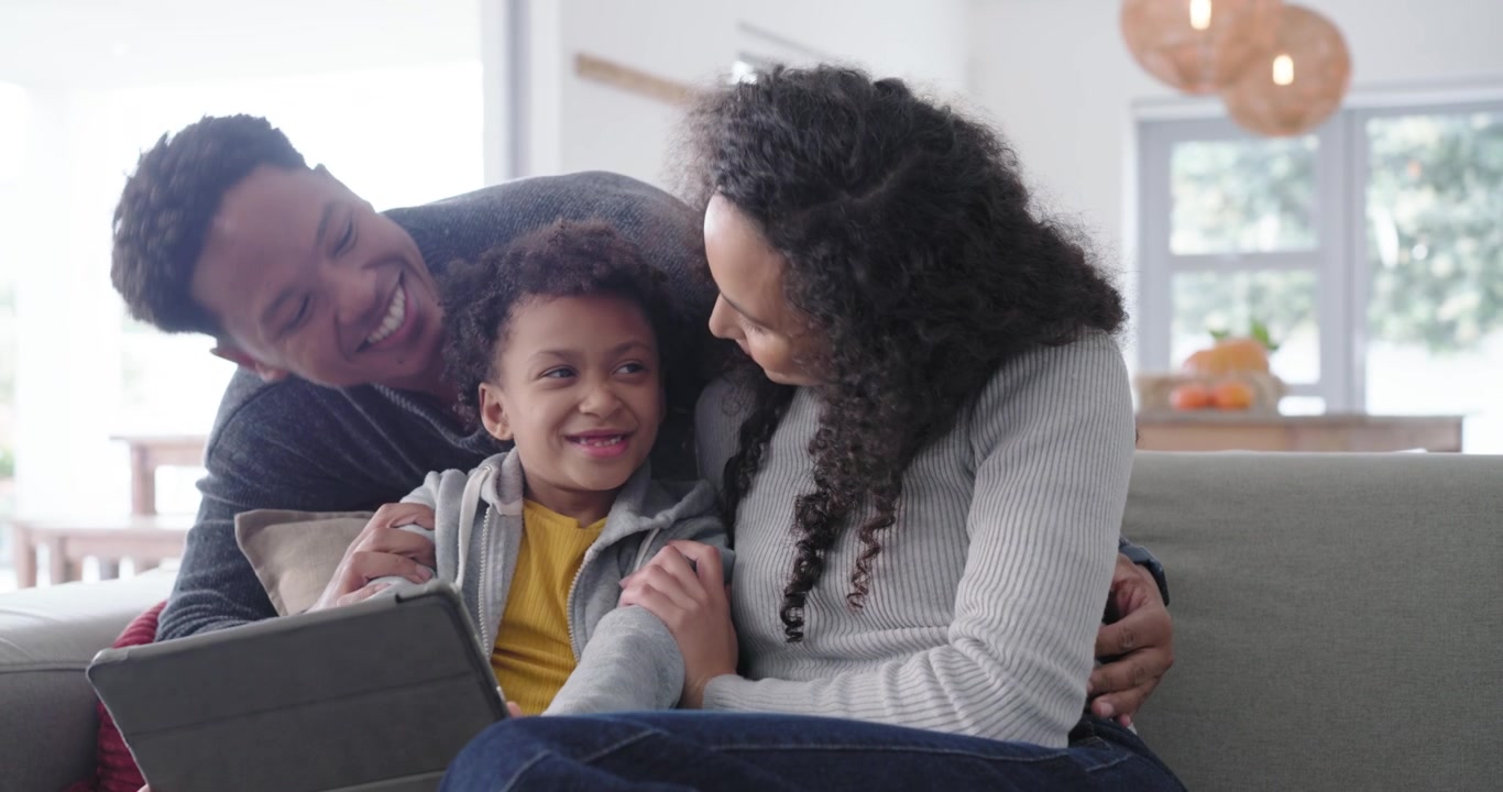 Famiglia afroamericana seduta sul divano a casa, bambino che tocca il tablet, genitori che si appoggiano e sorridono