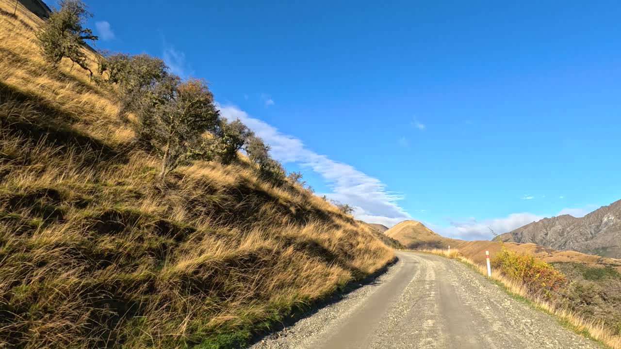 Vehicle travels winding gravel road, sunlit hills, blue sky, steady forward camera movement, natural scenery