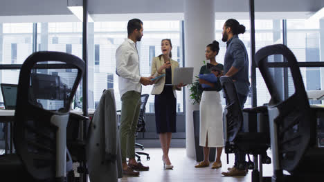 Mujer-De-Negocios-Caucásica-Sosteniendo-Una-Computadora-Portátil-Hablando-Con-Diversos-Colegas-De-Pie-En-Una-Reunión-De-Oficina