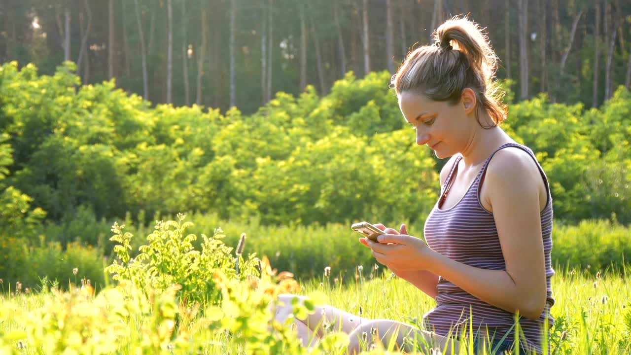 Happy girl sitting on green lawn and uses smartphone on Scenic Field at Sunset Background