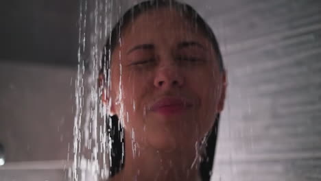 happy woman with water pouring down face in shower closeup. positive lady with closed eyes enjoys spa procedures in bathroom. hygienic routine