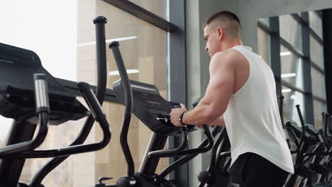 Side view of gym junkie on elliptical trainer gripping handles and staring at console, muscles engaged during intense cardio session in bright urban fitness studio with glass windows and equipment