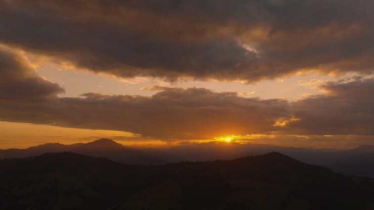 Aerial view showing an orange sunset as it casts a warm glow between a cloudy sky and distant mountain ranges