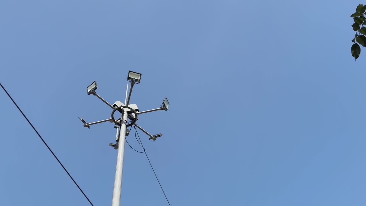 A tall floodlight tower with multiple LED lamps extends into a clear blue sky, captured from a low angle with minimal surroundings and clean, open space