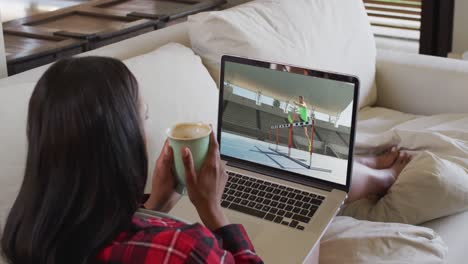 Composite-of-woman-sitting-at-home-holding-coffee-watching-athletics-running-event-on-laptop