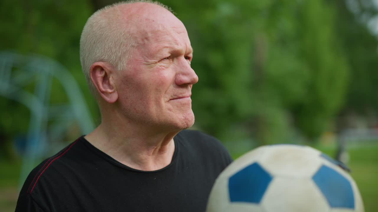 An elderly man rolls a soccer ball in his hand with a focused expression, with a blur view of trees and other equipment in the background