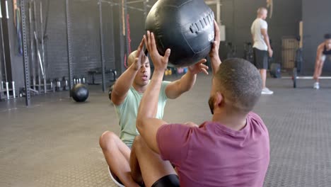 Dos-Hombres-Diversos-Haciendo-Ejercicio-Juntos,-Haciendo-Abdominales-Y-Pasando-Pelota-Medicinal-En-El-Gimnasio,-En-Cámara-Lenta