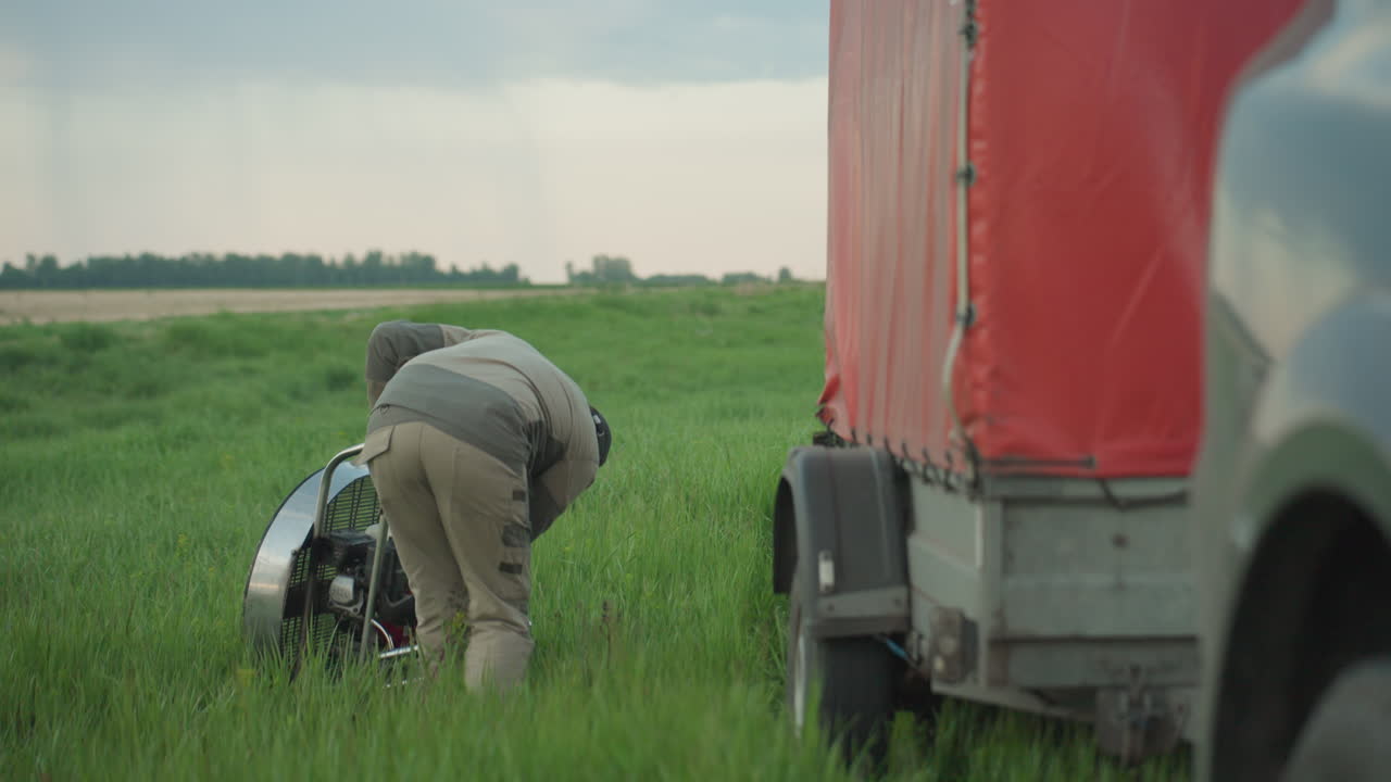 close up of man adjusting paramotor engine by circular fan cage mounted on wheeled trailer in lush green field at dusk, focus on precise hand movement