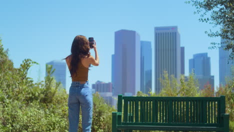 unknown woman standing in park making photo skyscrapers on smartphone back view.