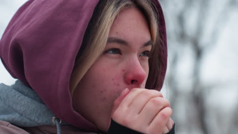 close-up of girl with spots on face standing in cold, rubbing nose and blowing hot air into hands to stay warm, hooded winter outfit emphasizes the freezing weather