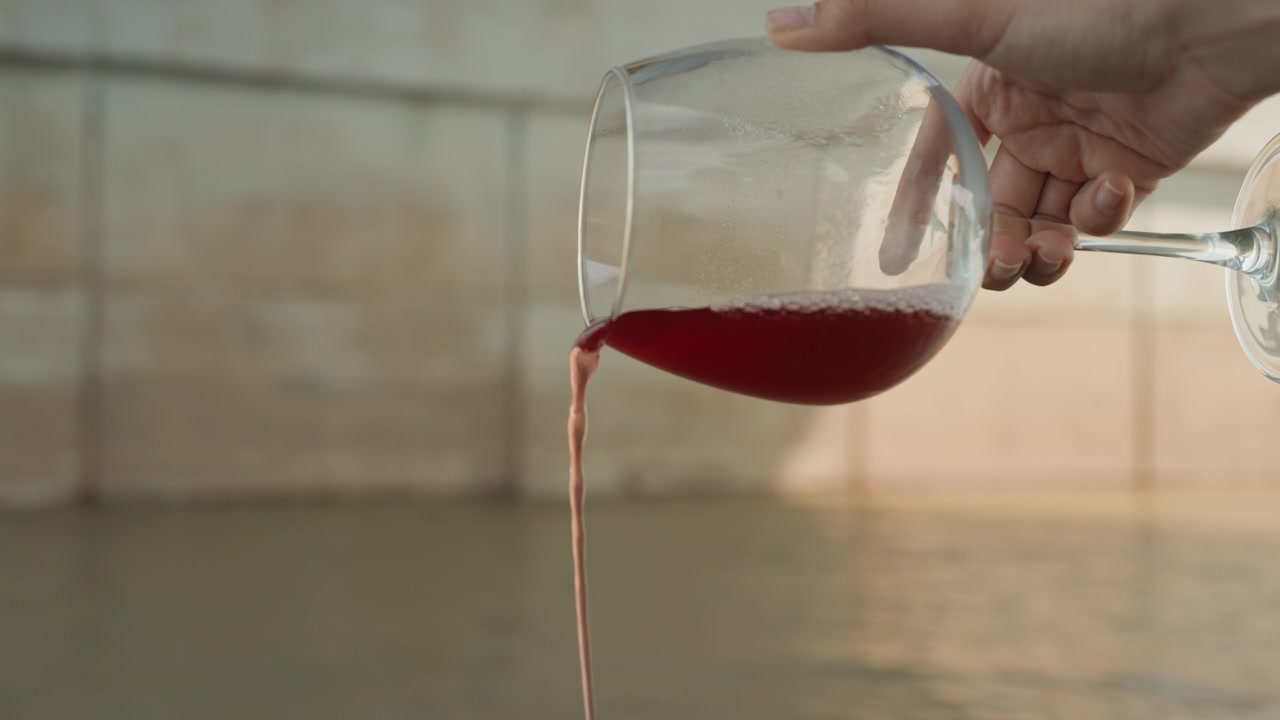 Una mano tiene un bicchiere da vino con liquido rosso, inclinandolo gradualmente per versarne il contenuto. Lo sfondo è illuminato delicatamente e sfocato, enfatizzando l'azione in una luce naturale soffusa.
