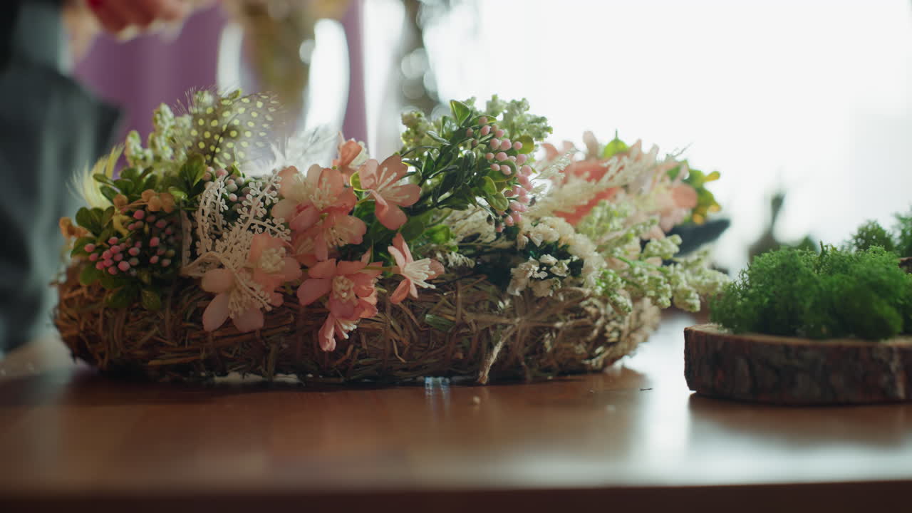 Female hand arranging floral wreath with pink blossoms, green leaves, and berries on wooden table near moss decorations, rustic handmade design illuminated by natural daylight in cozy creative workspace