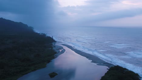 Aerial-view-of-waves-crashing-onto-the-shore-with-green-mountains-on-the-left,-set-against-a-dusky-sky-with-clouds-in-Colombia