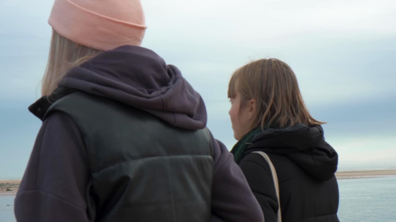 A young woman and a friend walk by the beach in Scotland, facing the water on a calm day