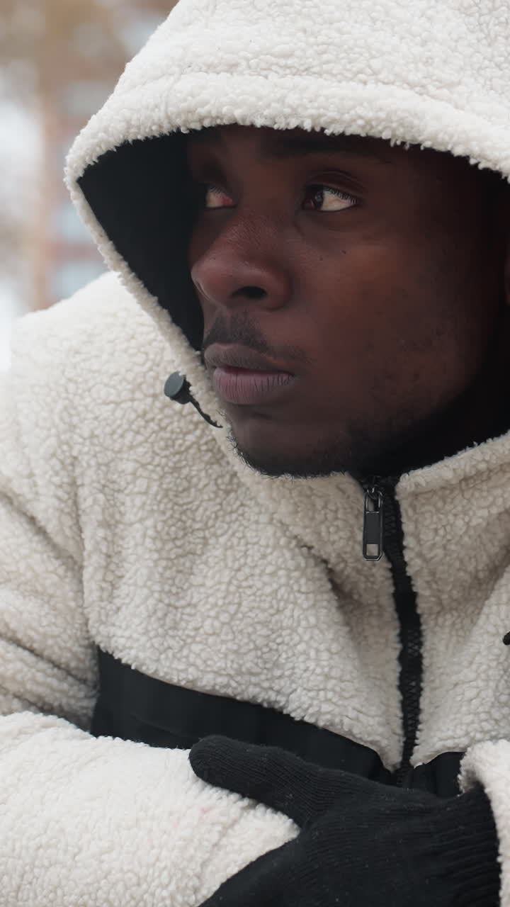 artista de rap con capucha blanca y guantes se apoya en una barra de hierro al aire libre, asintiendo con la cabeza en pensamiento, el fondo presenta terreno nevado, árboles y edificios distantes