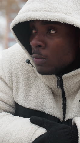 artista de rap con capucha blanca y guantes se apoya en una barra de hierro al aire libre, asintiendo con la cabeza en pensamiento, el fondo presenta terreno nevado, árboles y edificios distantes