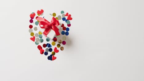 Video-of-white-gift-box-tied-with-red-ribbon-on-confetti-in-heart-shape,-with-copy-space