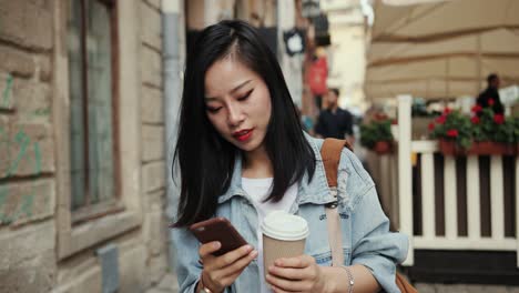 Feliz-Y-Atractiva-Mujer-Joven-Con-Estilo-Caminando-Por-La-Calle-Con-Un-Café-Para-Llevar-Y-Escuchando-El-Mensaje-De-Voz-De-Audio-En-El-Teléfono-Inteligente