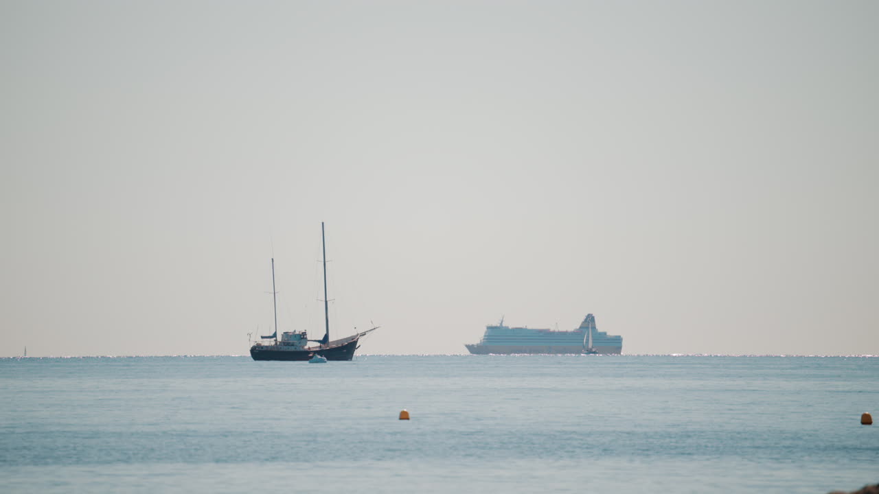 Wide shot of a calm sea with a sailboat and a large cruise ship sailing in the distance under a pale blue sky