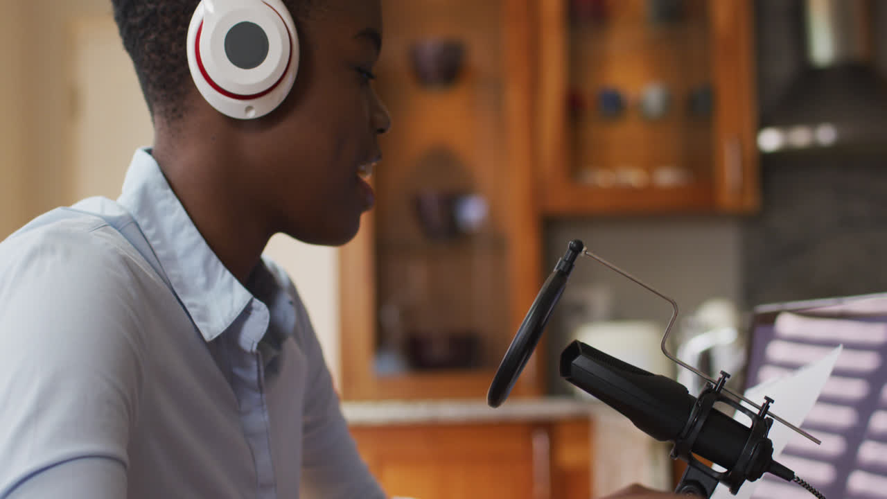 African american woman wearing headphones singing in microphone at home