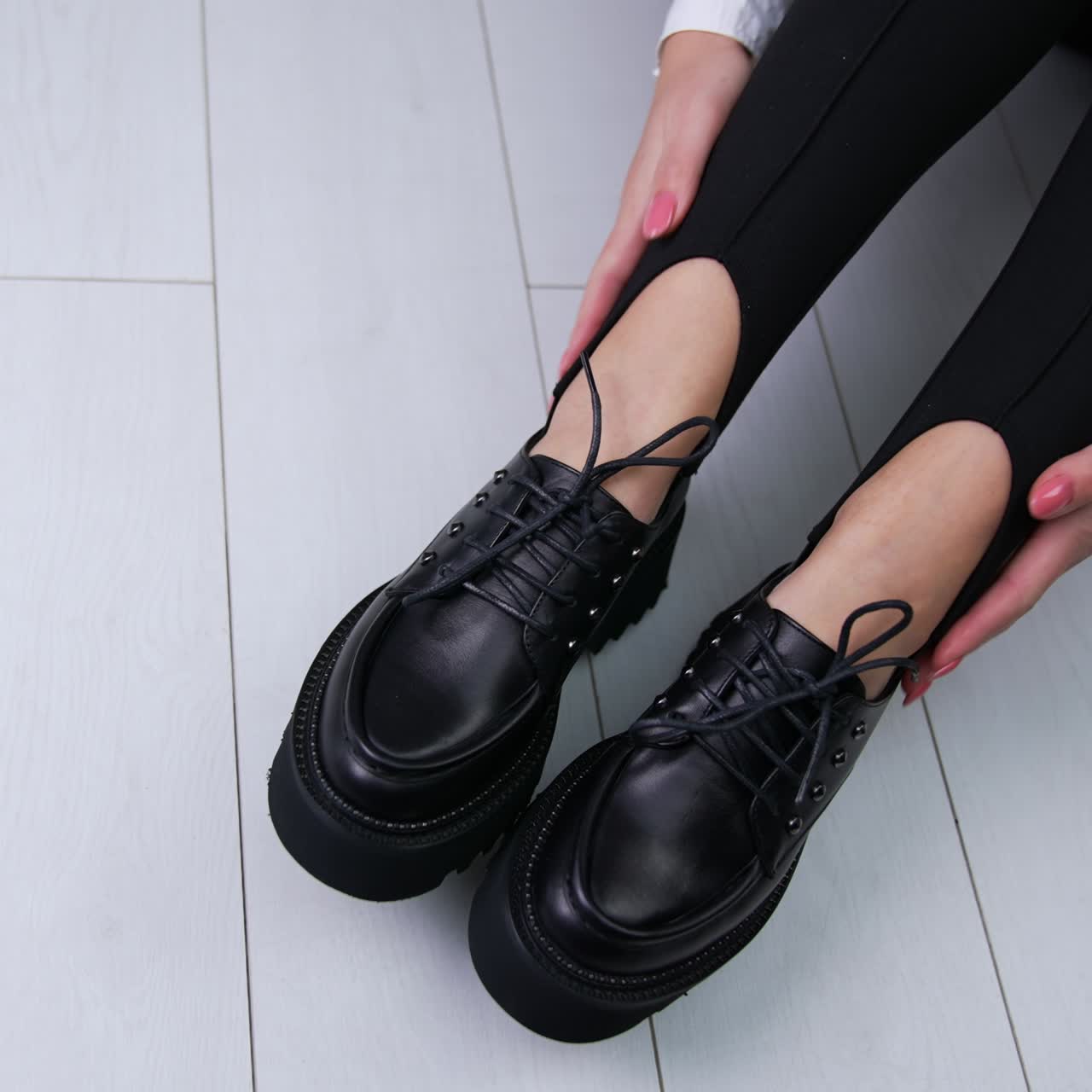 Lady demonstrates black shoes with laces sitting on the white floor. Woman touches her modern footwear and turns in front of camera. Close up. Top view
