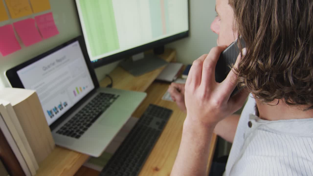 hombre caucásico enfocado trabajando remotamente, usando computadora portátil y hablando en teléfono inteligente