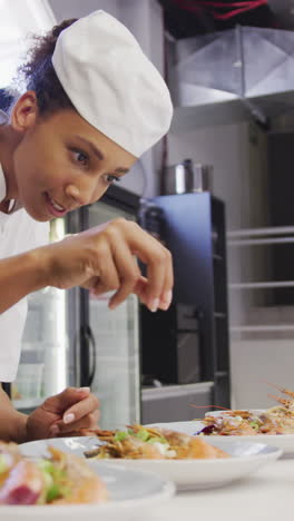 african american female chef wearing chefs whites in a restaurant kitchen, putting food on a plate