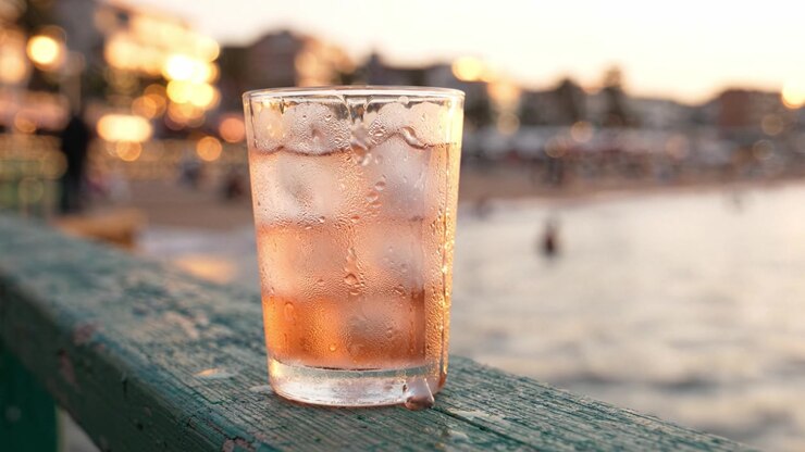 A refreshing glass of iced beverage sits on a wooden railing by the beach, capturing the warm glow of the sunset and the serene waves in the background.